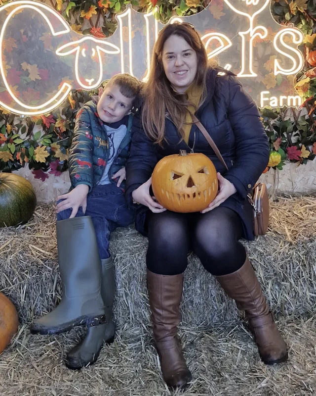 Laura and her son sitting on hay bales at a pumpkin patch, Laura holding a carved pumpkin, both smiling.